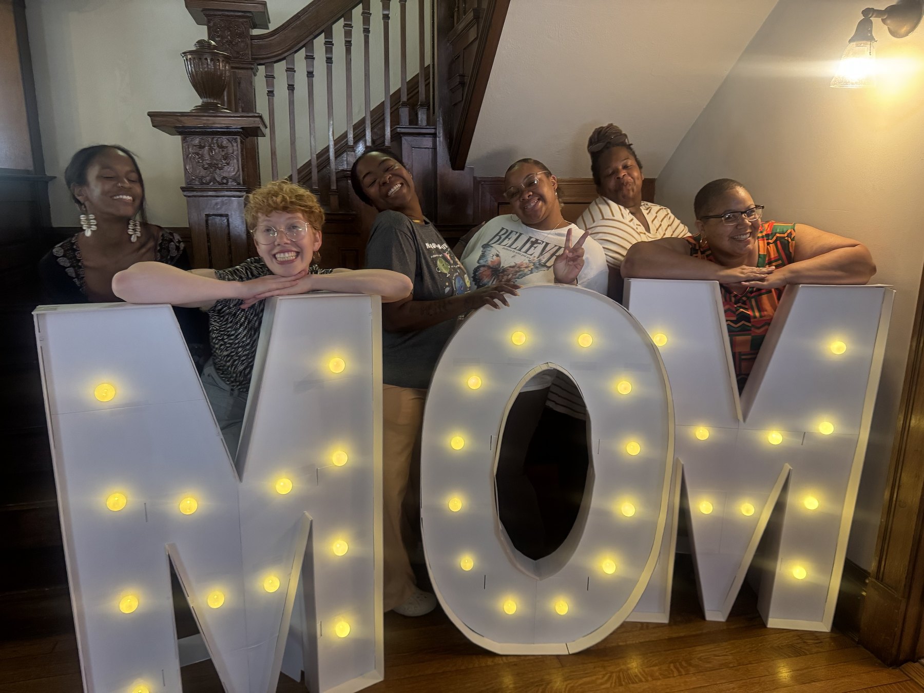 Six women laughing together behind a lit MOM marquee on the wooden staircase at Korédé House